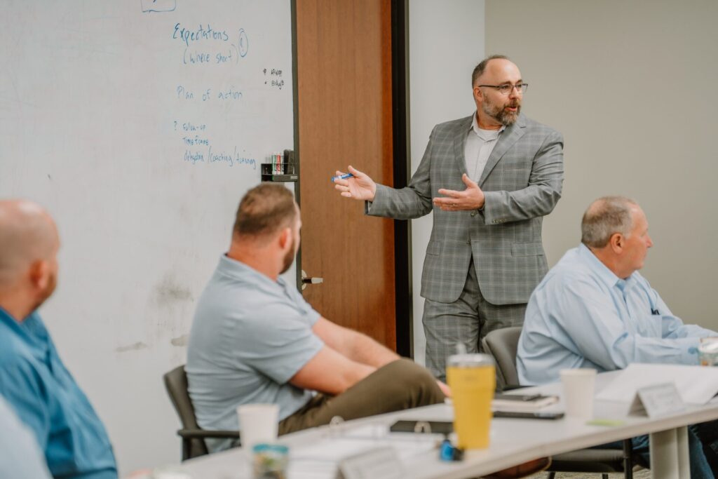 A man in a gray checked suit gestures while speaking to a group in a meeting room, imparting insights from executive training. Three seated men listen attentively. The man stands beside a whiteboard with handwritten notes. A door is partially visible in the background.