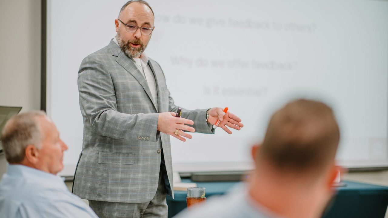 A man in a gray plaid suit is standing and gesturing while speaking to a group of seated individuals in a classroom or meeting room. A presentation slide with indistinct text is projected in the background. Attendees are listening attentively, engaged in leadership training.