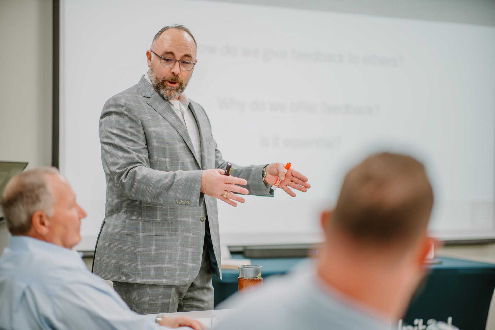 A man in a gray plaid suit is standing and gesturing while speaking to a group of seated individuals in a classroom or meeting room. A presentation slide with indistinct text is projected in the background. Attendees are listening attentively, engaged in leadership training.