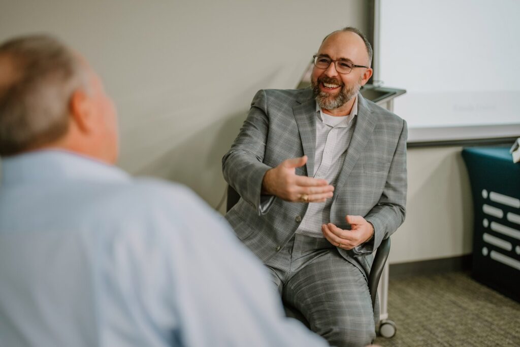 2023-01-18 Rmada-159 A man wearing a gray checkered suit and glasses is smiling and extending his hand to another person. He appears to be engaged in a friendly conversation about executive training in an office setting. The second person is slightly blurred in the foreground, dressed in a light-colored shirt.