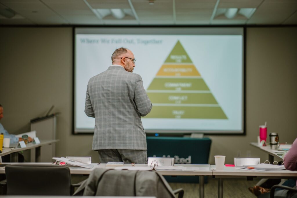 A man in a gray plaid suit stands facing a large presentation screen displaying a pyramid chart labeled with tiers such as Trust, Conflict, Commitment, Accountability, and Results. The room is set up for executive training with rows of tables scattered with papers and cups.