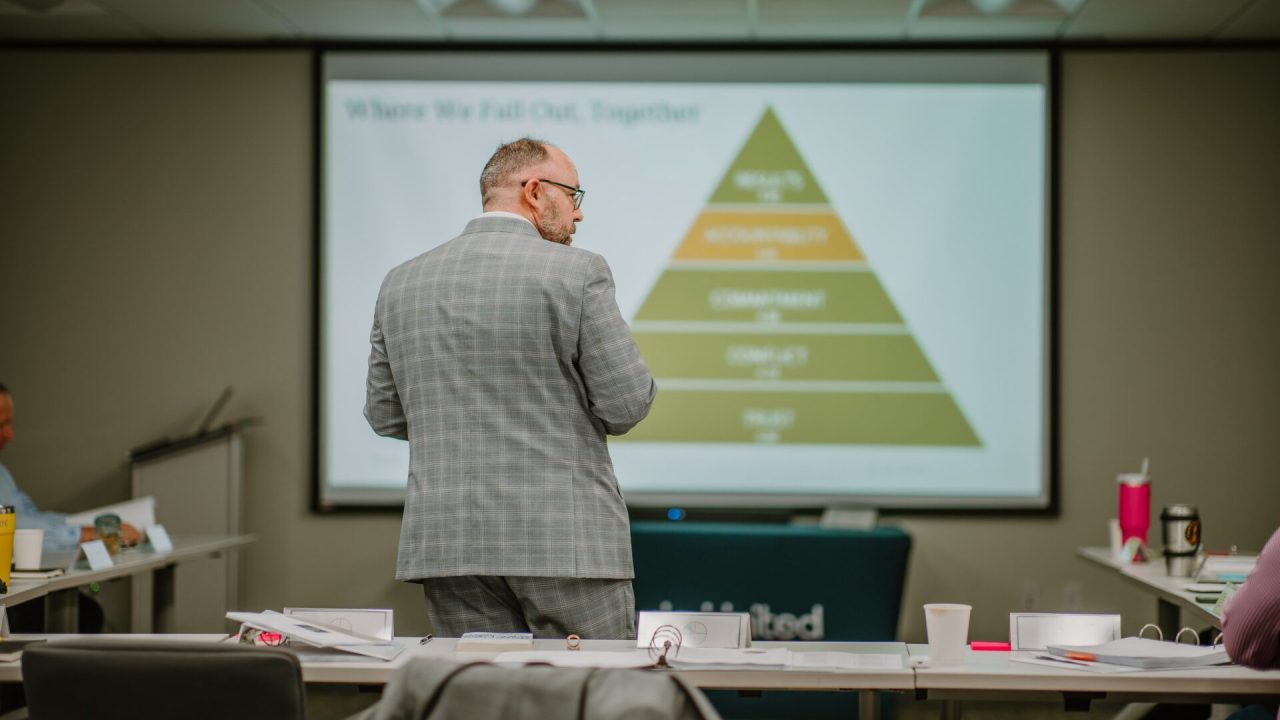 A man in a gray plaid suit stands facing a large presentation screen displaying a pyramid chart labeled with tiers such as Trust, Conflict, Commitment, Accountability, and Results. The room is set up for executive training with rows of tables scattered with papers and cups.