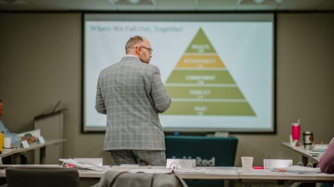 2023-01-18 Rmada-205 A man in a gray plaid suit stands facing a large presentation screen displaying a pyramid chart labeled with tiers such as Trust, Conflict, Commitment, Accountability, and Results. The room is set up for executive training with rows of tables scattered with papers and cups.