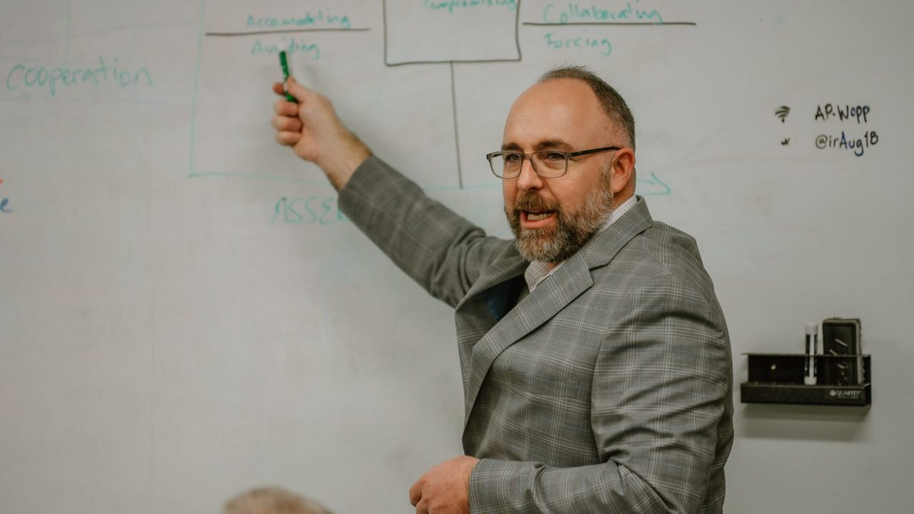 A man in a grey suit gestures with a green marker towards a whiteboard covered with diagrams and text. He appears to be explaining something during an executive training session. There is a partially visible person seated in the foreground, and markers are held in a tray on the wall.