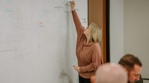 2023-01-18 Rmada-263 A person with blonde hair, wearing a brown sweater, stands near a whiteboard using a marker to write or draw. The whiteboard contains various illegible markings from the ongoing leadership training session. A few people are seated, facing the whiteboard, with the back of one person's head visible in the foreground.