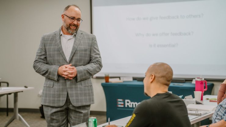 2023-01-18 Rmada-99 A man in a gray plaid suit, wearing glasses, stands and speaks to a seated, attentive group in a classroom setting. Behind him, a screen displays a presentation slide titled "Feedback, Giving." Desks, notepads, and drinks are visible in the room during this executive training session.