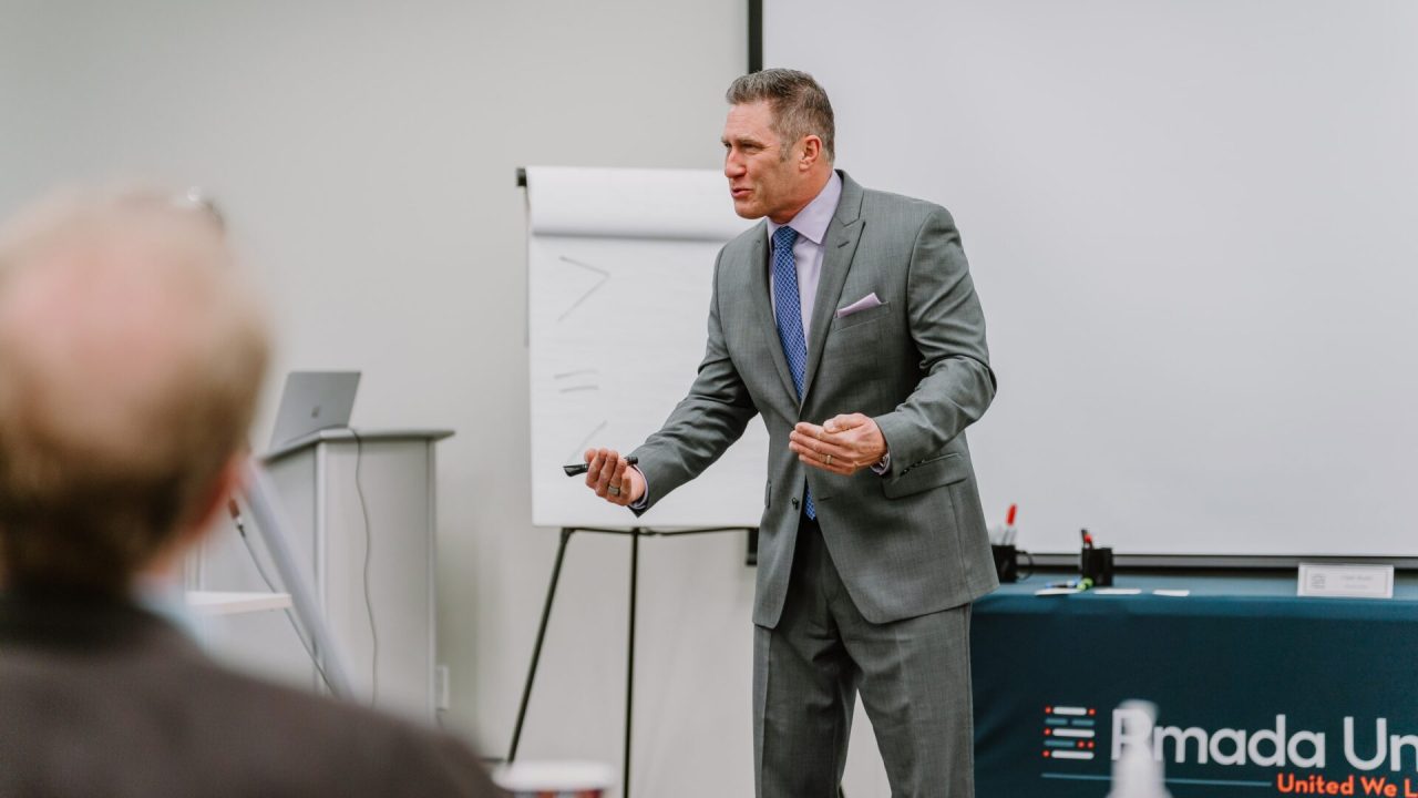 2023-03-22 Rmada-104 A man in a grey suit stands in front of a flip chart, holding a marker and speaking passionately. Behind him, a table with a "Ramada Un..." logo is partially visible. He appears to be leading an executive training session in a professional setting.