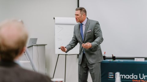 2023-03-22 Rmada-104 A man in a grey suit stands in front of a flip chart, holding a marker and speaking passionately. Behind him, a table with a "Ramada Un..." logo is partially visible. He appears to be leading an executive training session in a professional setting.
