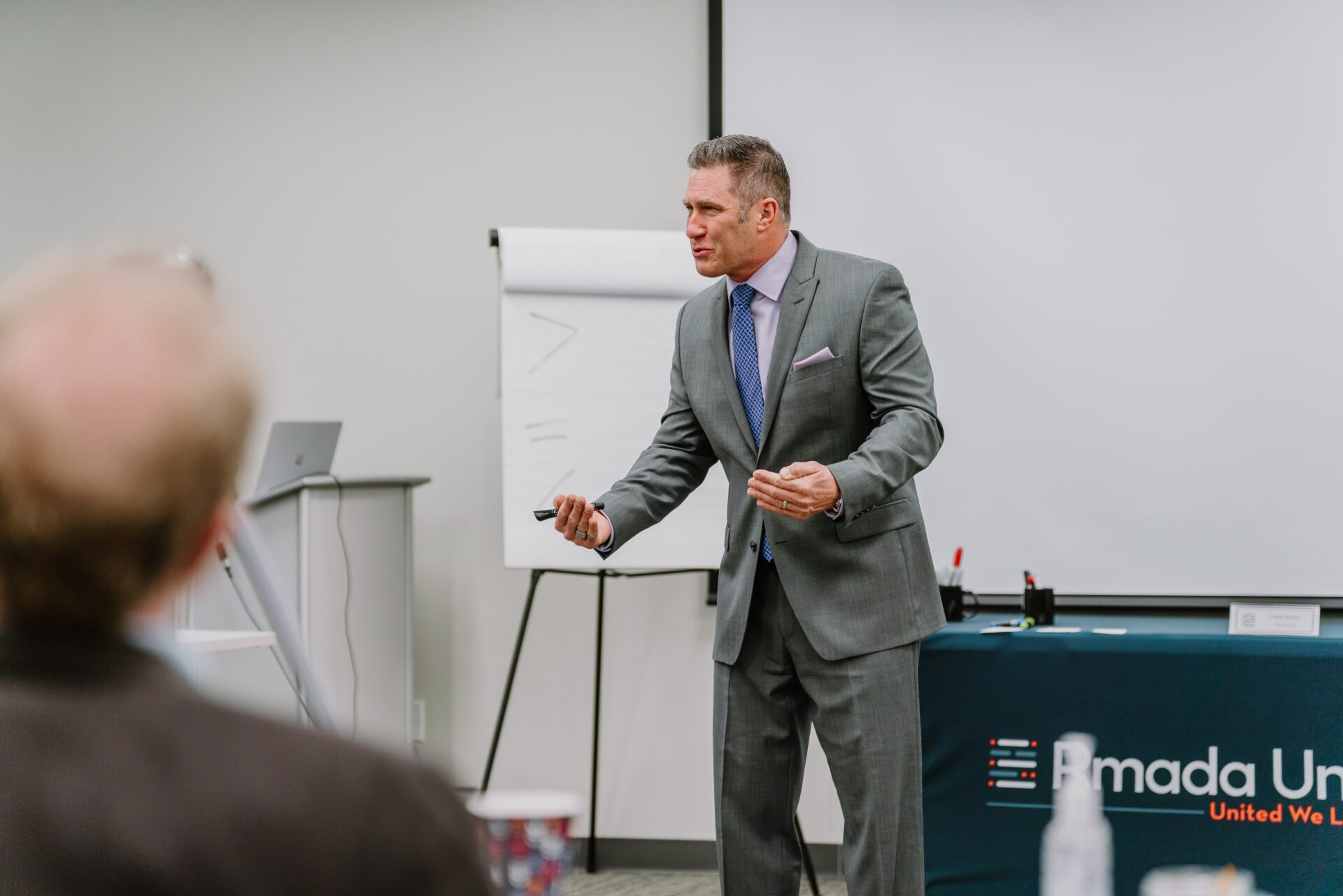 A man in a grey suit stands in front of a flip chart, holding a marker and speaking passionately. Behind him, a table with a "Ramada Un..." logo is partially visible. He appears to be leading an executive training session in a professional setting.