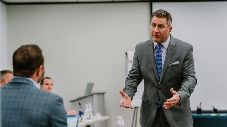 2023-03-22 Rmada-115 A man in a suit stands addressing a colleague in a meeting room, demonstrating the techniques from veteran leadership training. The man gestures with his hands while speaking to several people who sit around a table with laptops, a water bottle, and papers. A flip chart and a screen are visible in the background.