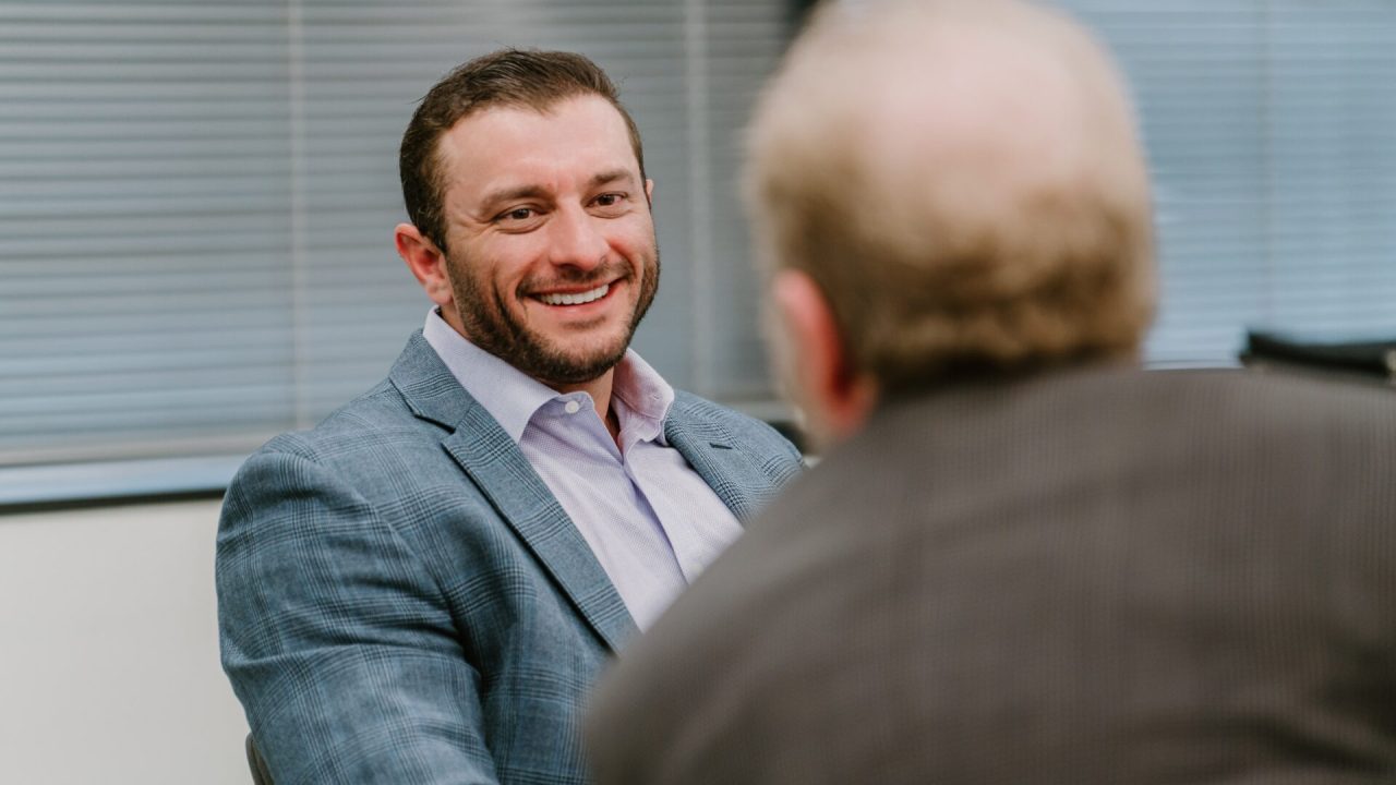 A man in a blue suit and white shirt is smiling during a conversation with another man whose back is to the camera. They are in an indoor office setting with blinds covering the windows, likely discussing veteran leadership training.