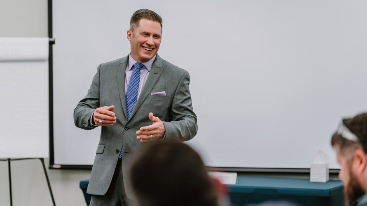 A man in a grey suit and purple tie stands in front of a whiteboard and a screen, speaking and gesturing to an unseen audience. He is smiling, appearing engaged and enthusiastic. Two people in the foreground are listening attentively at this executive training session.