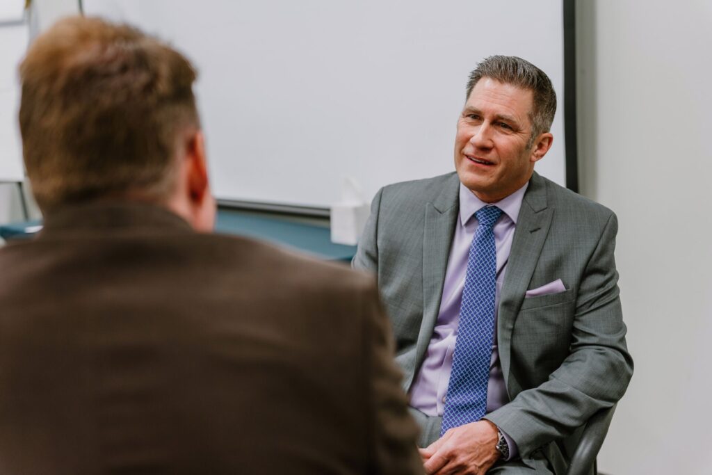 2023-03-22 Rmada-43 Two men are having a discussion in an office setting. One in a suit and tie sits facing the other, who is only partially visible from the back. The background shows a whiteboard and office furnishings, suggesting an environment focused on executive training. The atmosphere appears professional and conversational.