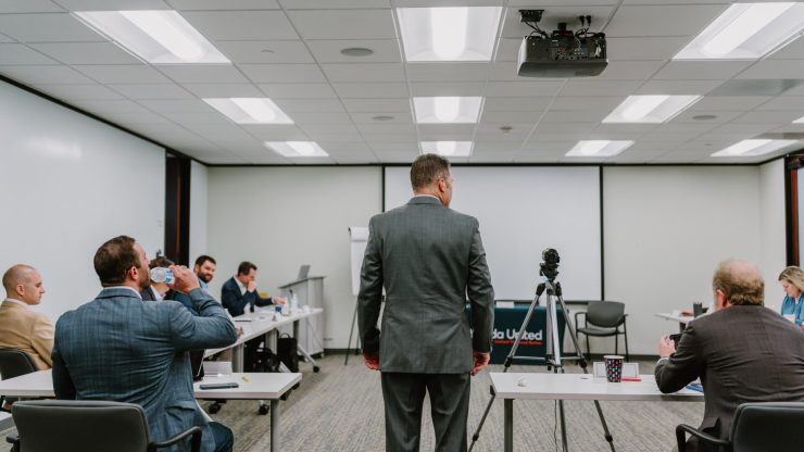 2023-03-22 Rmada-60 A group of people are seated at tables in a conference room for executive training. A man stands facing them, positioned near a tripod with a camera. The room has white walls, a projector, and a ceiling with fluorescent lights. Some attendees are engaged in discussion, while others are drinking water.