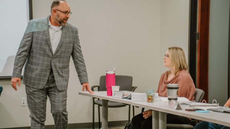 A man in a gray checked suit stands and speaks to a woman seated at a table during an executive training meeting in the office. The woman, wearing a pink sweater, has a pink tumbler and other items on the table in front of her. Other participants are partially visible.