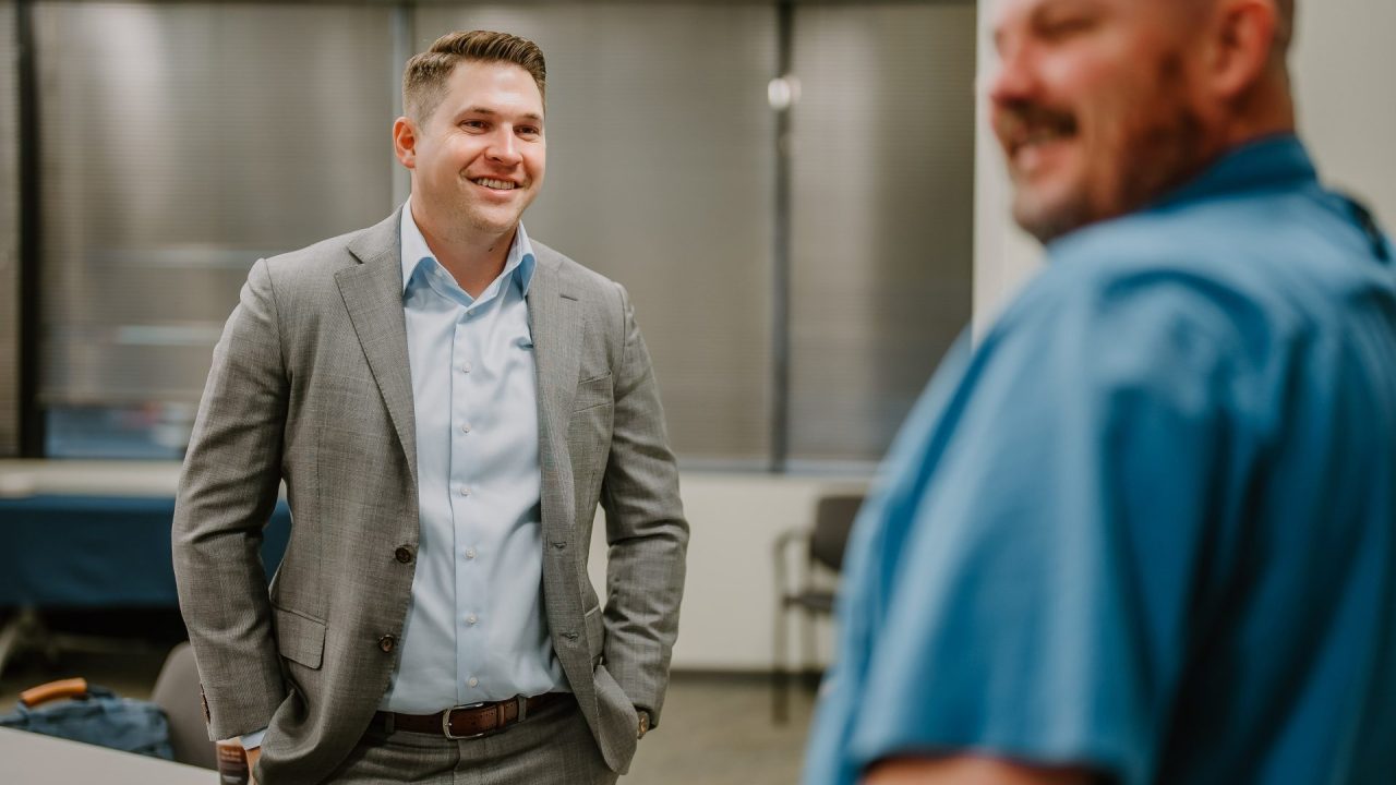 2023-01-18 Rmada-154 A man in a grey suit is standing and smiling while conversing with another man in a blue shirt who is slightly out of focus. They are in an office setting with blinds covering the windows in the background, likely discussing executive training. The atmosphere appears friendly and informal.