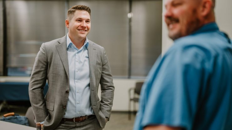 A man in a grey suit is standing and smiling while conversing with another man in a blue shirt who is slightly out of focus. They are in an office setting with blinds covering the windows in the background, likely discussing executive training. The atmosphere appears friendly and informal.