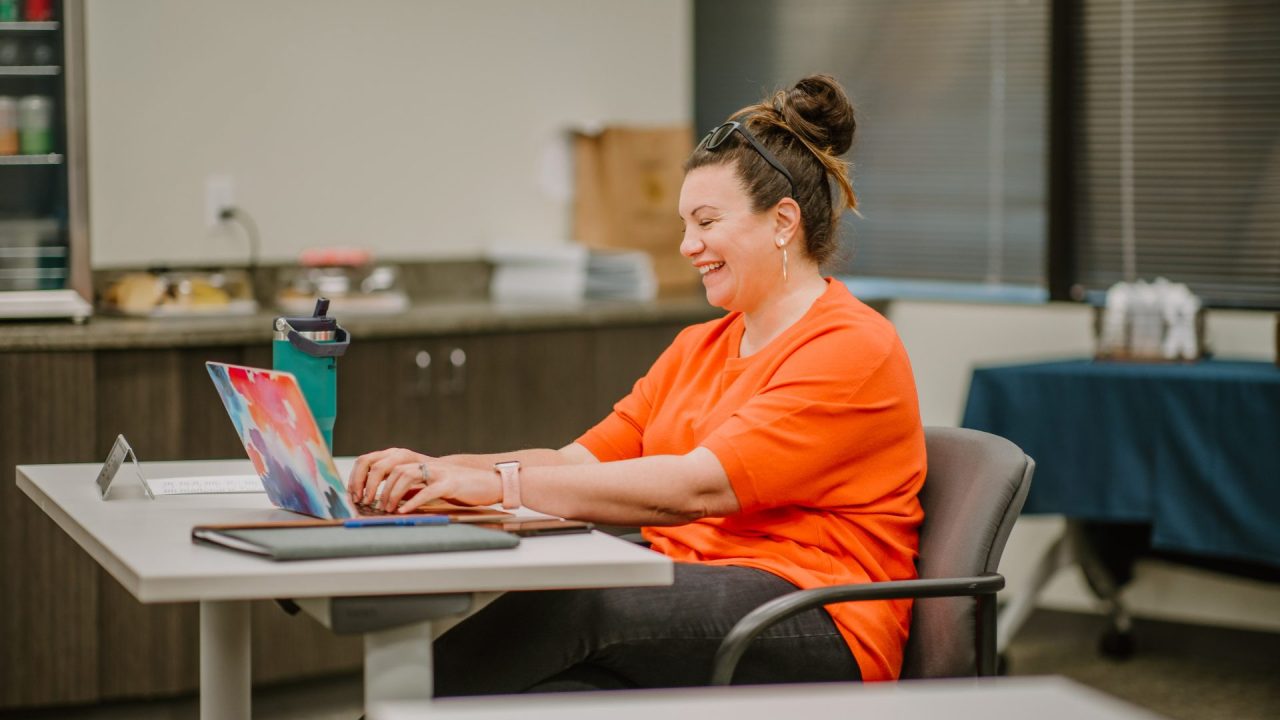 A woman with a bun, wearing an orange top, is sitting at a desk and smiling while typing on a colorful laptop. She has a smartwatch on her wrist and a water bottle next to her. The background shows an office space with cabinets and paperwork from an ongoing executive training session.