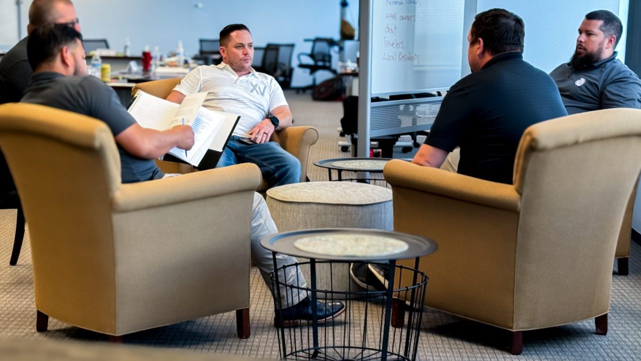 rmada Four men are seated in armchairs, engaged in a discussion. One man is holding an open binder, while the others listen attentively. There is a flip chart in the background with some partially visible writing, setting the scene for what appears to be an executive training session in a casual meeting room.